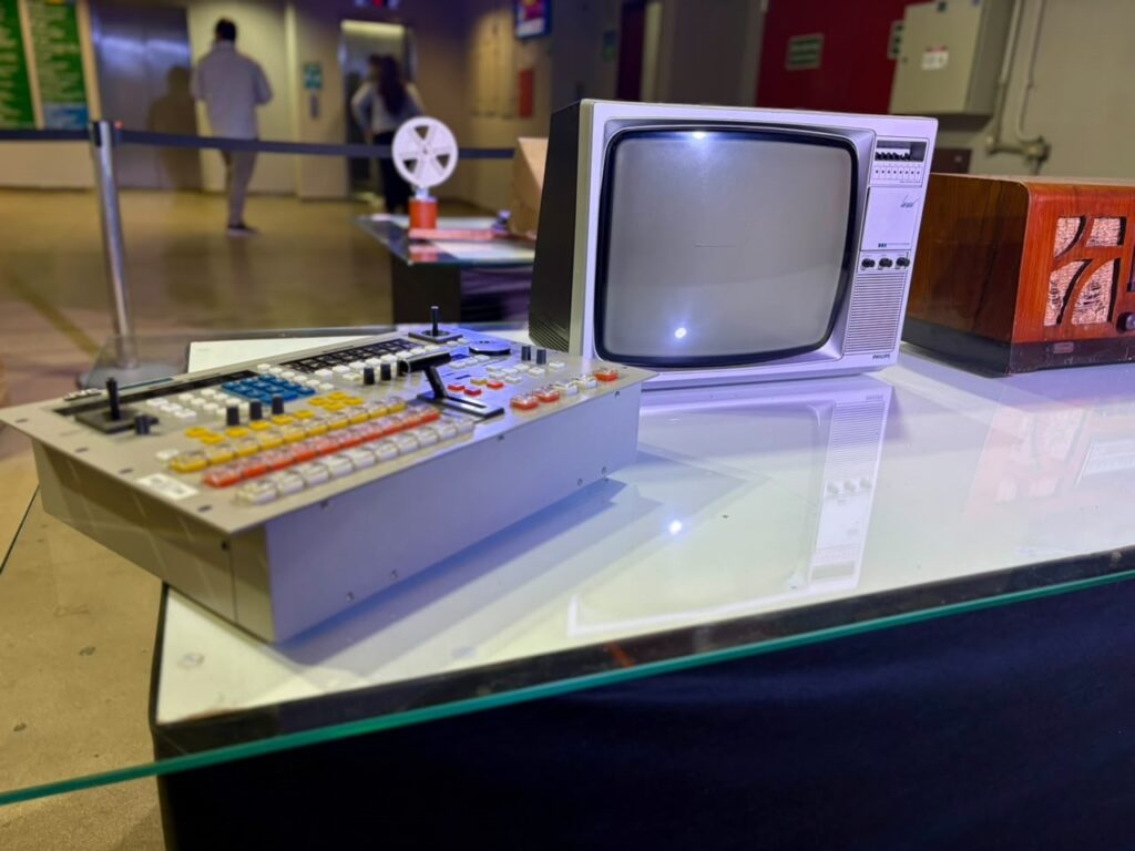 Esta é uma fotografia tirada em um ambiente que parece ser uma exposição cultural ou museu sobre a história da comunicação. O foco está em uma mesa de vidro onde estão expostos equipamentos de áudio e vídeo vintage: Em primeiro plano: Uma mesa de corte de vídeo (switcher) profissional, na cor cinza, repleta de botões coloridos (amarelo, laranja, azul e branco) e alavancas. É um equipamento clássico de estúdios de televisão de épocas passadas. À direita da mesa: Um televisor de tubo (CRT) da marca Philips, com um design típico dos anos 80 ou 90, com botões de controle na parte frontal. Ainda à direita: Parte de um rádio antigo de madeira, com detalhes entalhados. Ao fundo: O ambiente é amplo e iluminado, com pessoas circulando. Há um suporte com um carretel de filme (estilo película de cinema) em cima de outra mesa, reforçando a temática nostálgica da exposição. É uma cena bem técnica e nostálgica, focada no equipamento que moldou a era analógica da TV.