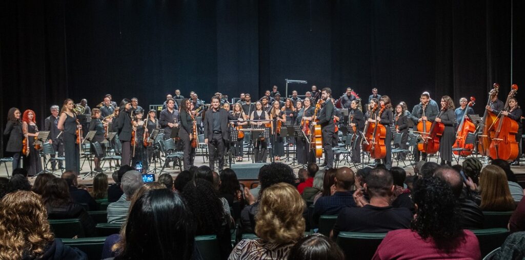 A imagem mostra uma orquestra sinfônica completa em pleno palco de um teatro, durante uma apresentação. Os músicos estão dispostos em semicírculo, com as cordas (violinos, violas, violoncelos e contrabaixos) ocupando a parte frontal e central, e outros instrumentos como sopros e percussão posicionados ao fundo. No centro do palco, um maestro está regendo o grupo. Todos os músicos estão vestidos formalmente, principalmente de preto, e estão sentados em cadeiras com estantes de partitura à frente. O palco é de madeira e o ambiente é iluminado de forma focada nos músicos, enquanto o fundo é escuro. Em primeiro plano, vê-se a plateia sentada em poltronas, observando a apresentação.