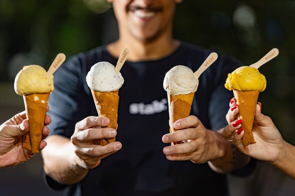 Nesta imagem, quatro mãos estão segurando casquinhas de sorvete em direção à câmera, criando uma cena convidativa e alegre. Aqui estão os detalhes: Os sorvetes: Cada casquinha contém uma bola de sorvete de sabor diferente, variando entre tons de amarelo, creme e bege, com texturas que sugerem sabores como frutas tropicais ou castanhas. Cada uma tem uma pequena espátula de madeira com a marca "Stab." gravada. As pessoas: No centro, levemente desfocado ao fundo, está uma pessoa vestindo uma camisa preta com a mesma marca "Stab." estampada, segurando duas das casquinhas. Nas laterais, outras duas pessoas (uma com unhas pintadas de vermelho vibrante) seguram as outras duas casquinhas. O ambiente: O foco está totalmente nos sorvetes, com o fundo desfocado, o que destaca o produto. A iluminação é quente e profissional, típica de uma foto promocional para uma sorveteria.