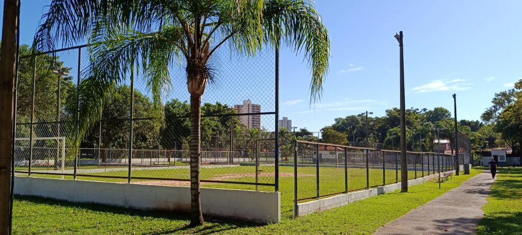 Esta foto mostra um campo de futebol ou área recreativa ao ar livre em um dia ensolarado. Aqui estão os detalhes principais: O Campo: É uma quadra cercada por grades metálicas, com uma superfície que mistura grama e terra batida. A estrutura parece ser dedicada a atividades esportivas. Elementos em destaque: Bem em primeiro plano, há uma palmeira alta que corta a visão do campo. Entorno: Ao redor do campo, há bastante gramado verde, árvores e, ao fundo, é possível ver alguns prédios, sugerindo que o local fica em uma área urbana. Caminho: À direita, há uma calçada de concreto que percorre o lado de fora da cerca, onde uma pessoa aparece caminhando sozinha ao longe. Iluminação: O céu está azul e limpo, indicando um dia claro e agradável. O cenário transmite uma sensação de tranquilidade, típico de praças ou parques públicos em bairros residenciais.