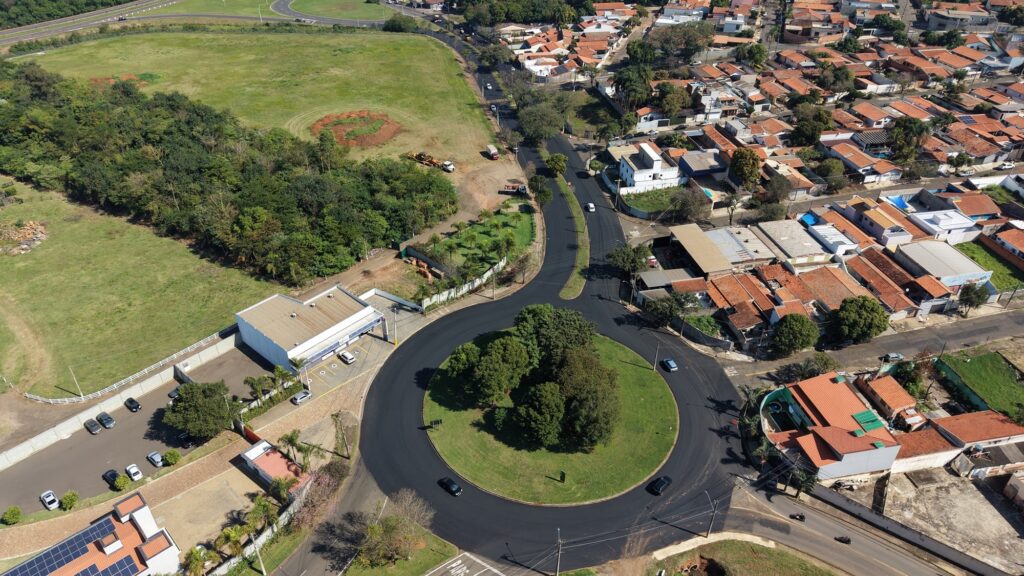 Esta é uma vista aérea impressionante de uma rotatória localizada em uma área urbana, com características bem distintas: A Rotatória e o Asfalto: O elemento central é uma grande rotatória com uma área verde no meio (árvores e gramado). O asfalto ao redor parece ser novo, bem escuro e liso, contrastando com as ruas ao redor. A Edificação Comercial: À esquerda da rotatória, há um edifício comercial com um telhado cinza, um estacionamento com alguns carros e uma área cercada. O Entorno: À esquerda, vê-se uma vasta área verde (campo/pasto) com algumas árvores e vegetação mais densa na divisa. À direita e na parte superior, existe um bairro residencial denso, caracterizado pelas casas com telhados de barro (tons avermelhados/alaranjados). É possível notar o fluxo de veículos circulando pela rotatória e pelas vias próximas. Iluminação e Clima: O dia está ensolarado e claro, com sombras bem definidas projetadas pelas árvores e edificações, indicando que a foto foi tirada provavelmente durante o período da manhã ou tarde.