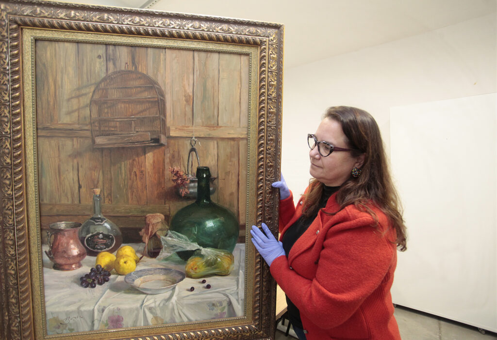 A imagem mostra uma mulher segurando com cuidado uma pintura a óleo emoldurada, provavelmente durante um processo de catalogação ou exposição.Detalhes da cena:Mulher: Está à direita, de perfil. Usa casaco vermelho sobre blusa preta, óculos de armação escura e brincos coloridos. Nas mãos, luvas de látex azuis, o que indica manuseio de acervo. Ela segura a lateral da moldura e olha atentamente para a obra.Quadro: É uma natureza-morta grande, com moldura dourada trabalhada e ornamentada. A pintura retrata uma mesa coberta por toalha branca com flores, sobre uma parede de madeira. Na mesa: um jarro de cobre, garrafa de vidro, limões, uvas, um prato, um garrafão verde grande, um pote e uma abóbora embalada em plástico. Ao fundo: uma gaiola de pássaro vazia pendurada na parede de madeira e um bule suspenso com flores. A assinatura "Bauer" aparece no canto inferior esquerdo.Ambiente: Sala clara, com paredes brancas e piso de concreto. Parece um ateliê, reserva técnica de museu ou espaço de restauração, pelo cuidado no manuseio e pela neutralidade do local.A obra transmite clima rústico e doméstico, típico de naturezas-mortas do início do século XX. O uso das luvas e a atenção da mulher reforçam o valor histórico e artístico da peça.