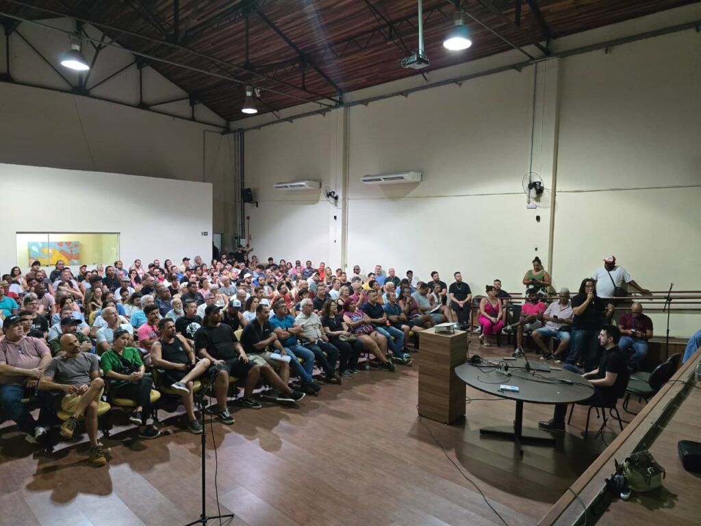Visão geral Auditório/salão interno com teto alto e estrutura metálica; piso de madeira. Espaço cheio: público numeroso sentado em fileiras voltadas para um pequeno palco/plataforma à direita da foto. Público Cerca de 120–160 pessoas (estimativa visual), de várias idades e gêneros; maioria sentada e atenta ao que acontece no palco. Pessoas conversam entre si e olham para frente; ninguém é identificável. Palco e oradores À direita, mesa redonda e uma pequena bancada/púlpito de madeira sobre o piso, com microfones e cabos; ao lado da mesa, duas ou três pessoas sentadas (uma falando ao microfone). Um microfone em pedestal no centro/primeiro plano aponta para a plateia, sugerindo espaço para perguntas ou interação. Ambiente e equipamentos Iluminação por lâmpadas pendentes no teto; ar‑condicionados de parede visíveis; ventilador montado na parede. Projetor suspenso no teto; cabos e equipamento de áudio próximos à mesa. Janela interna/vidro ao fundo esquerdo mostra um mural colorido em outra sala. Composição e sensação Foto tirada de um ângulo lateral elevado, mostrando a densidade do público e a relação entre plateia e oradores. Clima de reunião comunitária, palestra ou audiência pública — tom sério e participativo, com muitos presentes engajados. Detalhes observáveis Pessoas em pé e sentadas nas laterais e ao fundo, ocupando praticamente todo o espaço disponível. Itens pessoais (bolsas, mochilas) e equipamentos de som próximos ao palco.