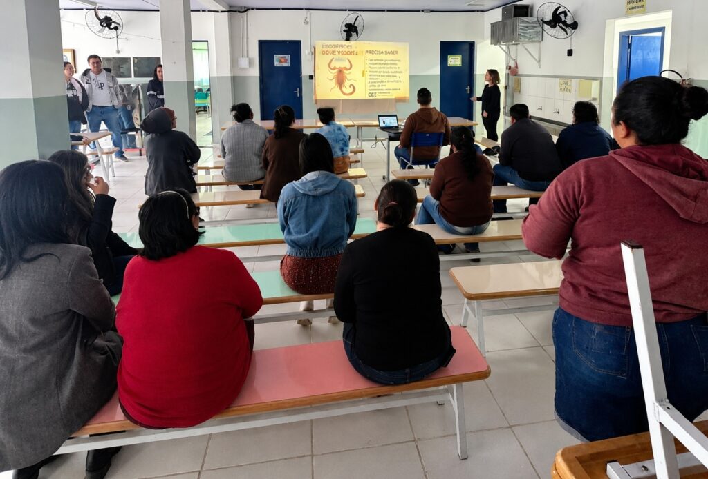 A imagem mostra uma palestra educativa ocorrendo em um auditório ou sala de espera. Há um grupo de pessoas sentadas em bancos de madeira (com assentos coloridos) viradas de costas para a câmera, assistindo a uma apresentação. Na frente, uma mulher está em pé, apresentando o conteúdo, e ao lado dela há um projetor exibindo um slide com o título "Escorpiões: O que você precisa saber", acompanhado por uma ilustração de um escorpião e um texto informativo. O ambiente é simples, com piso de cerâmica, paredes brancas e ventiladores fixados nas paredes e no teto.
