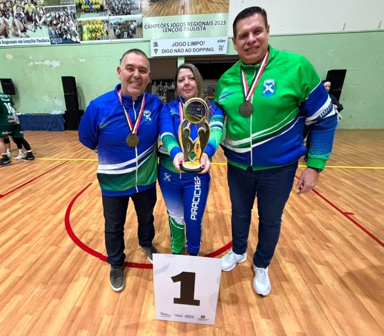 Esta é uma foto que celebra uma vitória esportiva. Três pessoas posam orgulhosas em uma quadra poliesportiva, indicando que conquistaram o primeiro lugar na competição. Aqui estão os detalhes: As Pessoas: Ao centro, uma mulher segura um troféu dourado. Ela está ladeada por dois homens; ambos usam medalhas penduradas no pescoço. Eles vestem uniformes esportivos coordenados nas cores azul, verde e branco. O Contexto: A placa com o número "1" no chão confirma a primeira colocação. Os banners ao fundo dizem "Campeões Jogos Regionais 2023 Lençóis Paulista" e reforçam a mensagem de "Jogo Limpo". Identificação: Na perna da calça da mulher, é possível ler "PIRACICABA", o que indica que eles estão representando a equipe desta cidade nos Jogos Regionais. É uma imagem que transmite muita satisfação pelo dever cumprido e pela conquista do pódio!