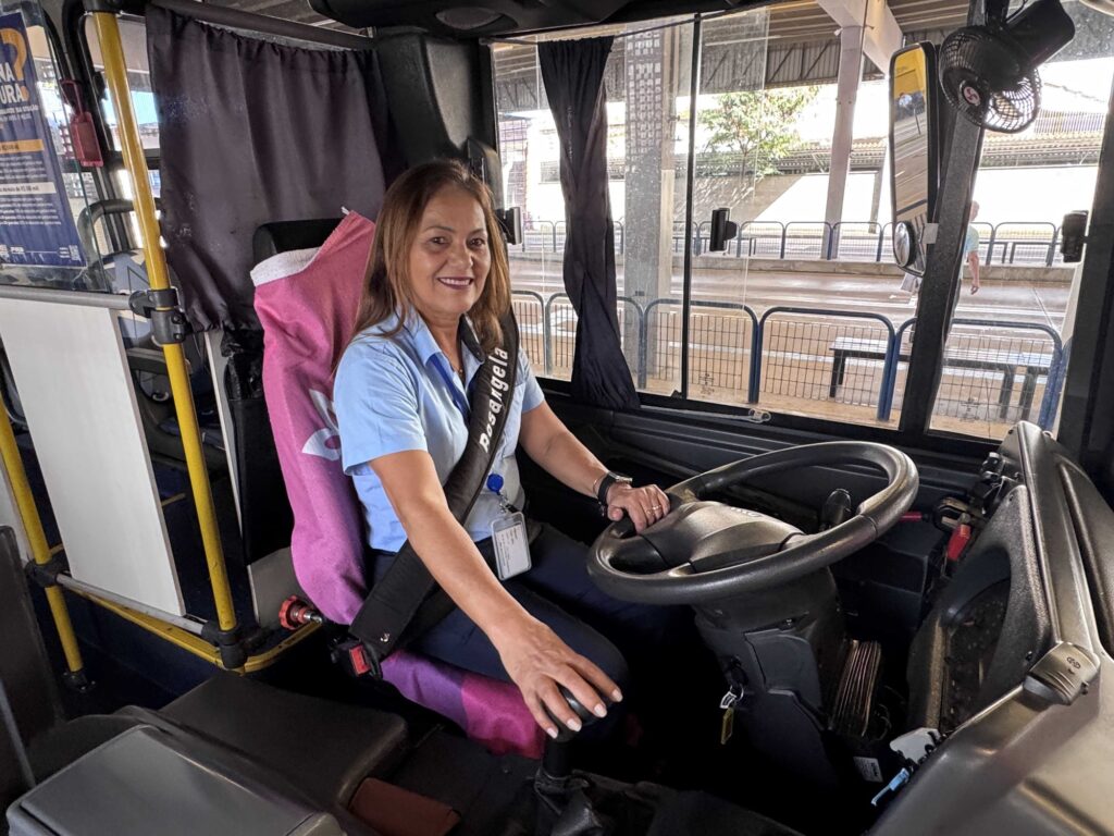 A imagem mostra uma motorista de ônibus sorridente, sentada ao volante no seu posto de trabalho. Aqui estão os detalhes: A pessoa: Uma mulher de cabelos castanhos, vestindo uma camisa polo azul claro. Ela está com uma faixa atravessada no peito onde se lê "Rosangela". O assento: A poltrona do motorista tem uma capa protetora rosa e preta encostada no encosto. O ambiente: Ela está dentro da cabine do motorista de um ônibus, com o volante à frente e um pequeno ventilador fixado na coluna da janela. O exterior: Através do para-brisa e das janelas laterais, é possível ver o ambiente externo, que parece ser um terminal de ônibus ou uma via urbana, com estruturas metálicas e grades de proteção. Há também um anúncio ou cartaz afixado na parte interna, próximo à janela esquerda.