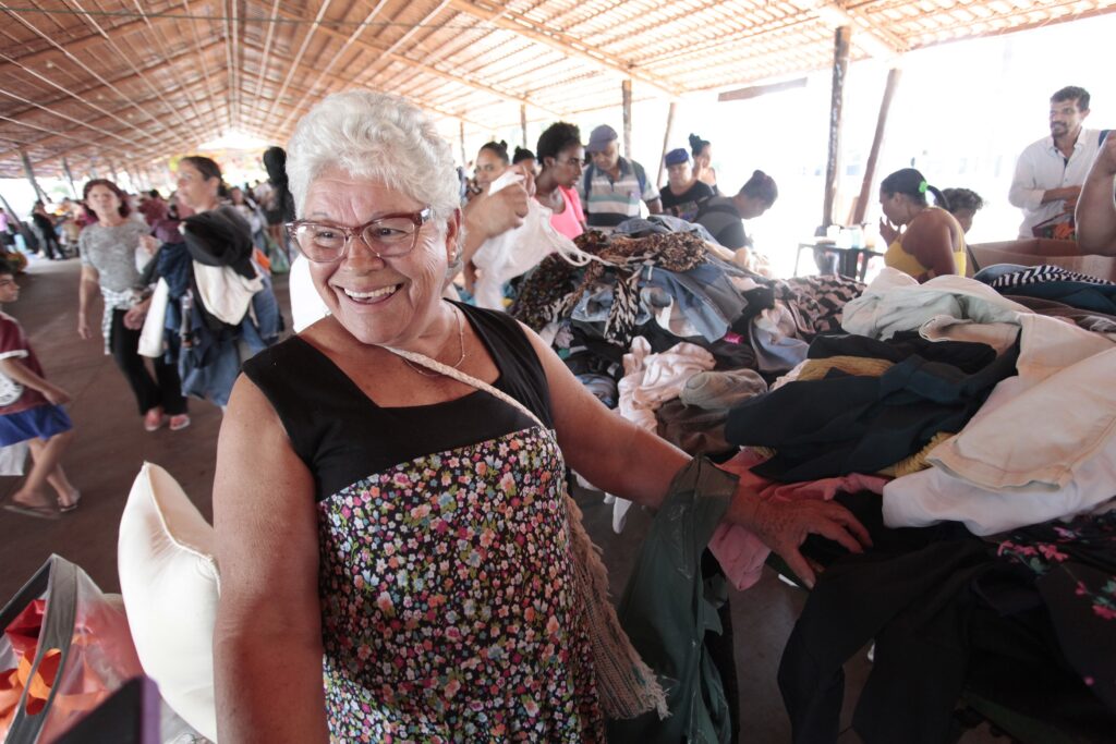Em primeiro plano, uma senhora de cabelos brancos curtos e óculos sorri alegremente para o lado. Ela usa uma blusa preta sem mangas sobre uma saia ou vestido com estampa floral colorida. Sua mão repousa sobre uma grande pilha de roupas dispostas em uma mesa. Ao fundo, em um espaço coberto com teto de palha, outras pessoas circulam e interagem com as peças de roupa, indicando um evento comunitário de distribuição ou troca.