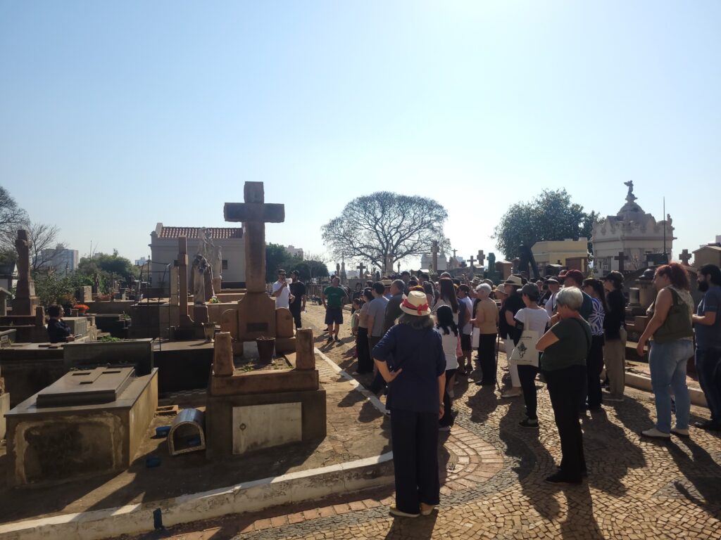 Foto panorâmica de um cemitério durante um evento ou visita guiada, com muitos visitantes reunidos. Observações principais: Local: cemitério com campas, mausoléus e cruzes de pedra em primeiro plano e ao fundo; arquitetura antiga visível em alguns túmulos. Pessoas: grande grupo de visitantes (idades variadas), muitos de pé formando uma roda/aglomerado à direita; alguns parecem ouvir um guia (um homem com microfone ou falando em direção ao grupo). Composição: cruz alta e escura em destaque à esquerda do centro; árvore grande sem muitas folhas ao centro-direita do horizonte; esculturas e detalhes ornamentais nos túmulos. Solo e textura: caminhos de paralelepípedo e achas de pedra, degraus e lajes nos túmulos; sombras longas projetadas pelas pessoas e pelas estruturas indicam sol forte e horário próximo ao meio-dia. Clima/iluminação: céu azul limpo, luz dura e direta, contraste alto entre áreas iluminadas e sombras. Atmosfera: tom respeitoso e contemplativo, mas também de interesse coletivo — aparente visita cultural, passeio histórico ou cerimônia comunitária.