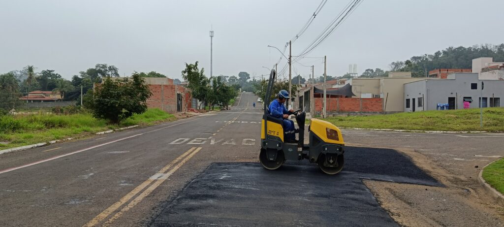Esta é uma imagem que registra um serviço de manutenção viária em andamento. Ação: Um operário, vestindo uniforme azul e capacete de segurança, está operando um rolo compactador vibratório amarelo (da marca Wacker Neuson). Ele está finalizando a compactação de um trecho de asfalto novo que foi aplicado recentemente no cruzamento. O Ambiente: É uma rua urbana pavimentada, com marcações de sinalização no solo, incluindo a palavra "DEVAGAR" pintada na via. O céu está nublado, indicando um dia típico de trabalho. Contexto: Ao fundo, vê-se uma paisagem urbana com casas, muros de tijolos aparentes, vegetação lateral e um terreno com uma subida ao longe. Há também postes de iluminação e fiação elétrica cruzando a cena. É uma foto que documenta o trabalho de reparo necessário para manter as condições de tráfego na via.