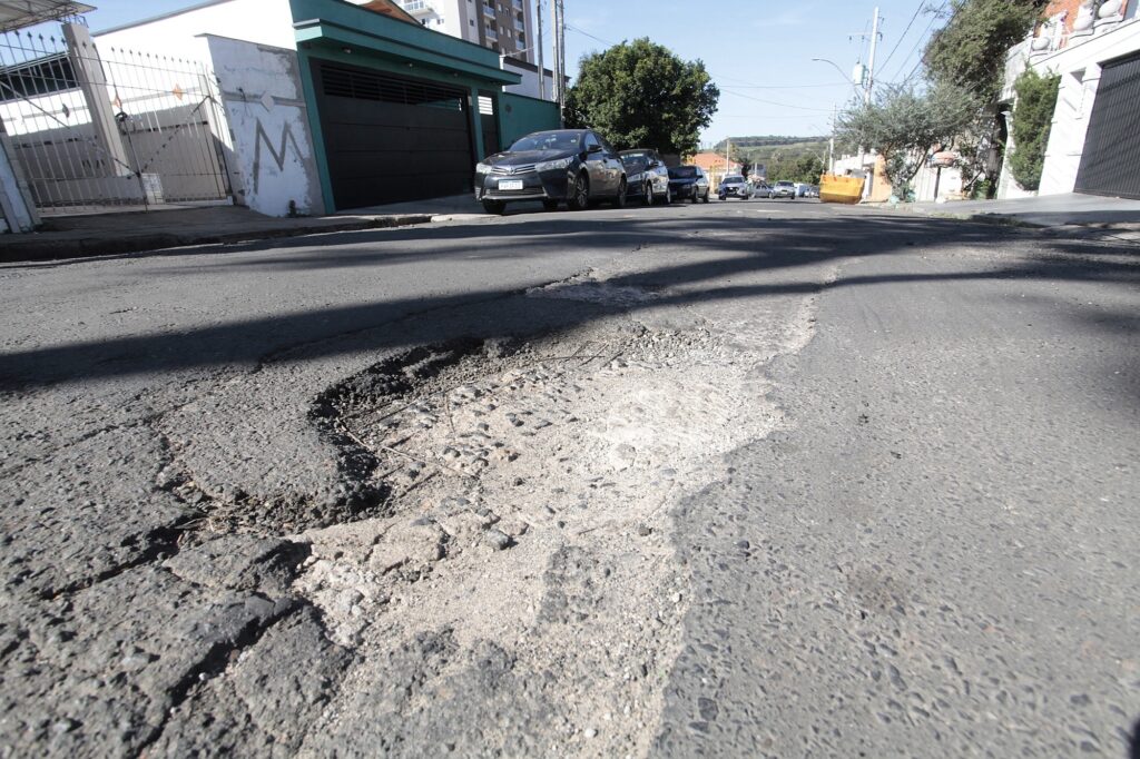 Imagem de uma rua inclinada com foco no asfalto danificado em primeiro plano. Primeiro plano: grande desgaste e buraco no pavimento; camada de base exposta e restos de brita solta, formando irregularidades perigosas para veículos e pedestres. Meio da cena: calçada e garagens residenciais à esquerda; um portão branco e uma fachada verde-azulada com porta de garagem escura. Vários carros estacionados ao longo da via à direita, alinhados morro acima. Vegetação: árvore de porte médio no meio-direita, com sombra projetada sobre a rua. Fundo: bairro residencial com postes e fiação, construções de pequeno e médio porte e horizonte com áreas verdes ao longe. Condição de luz: dia claro, céu azul e sombras bem definidas, indicando sol forte. A imagem transmite risco de dano a veículos e perigo para quem passa a pé por causa do pavimento comprometido.