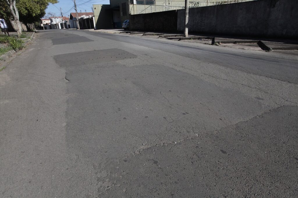 Rua residencial em leve subida com foco no pavimento desgastado. Pavimento: asfalto irregular com remendos recentes e manchas escuras; várias fissuras e pequenas depressões ao longo da pista. Em primeiro plano nota‑se uma área remendada retangular e trechos com desgate que podem causar desconforto a veículos. Meio da cena: sarjetas e boca de lobo ao longo da calçada direita, com trechos de meio‑fio quebrado ou rebaixado; faixa central da rua com desníveis visíveis. Laterais: à direita, muro alto com cerca‑concertina no topo; poste de concreto sobre a calçada. À esquerda, árvores e faixa de grama entre calçada e via; ao fundo, casas e garagens. Tráfego e segurança: vários carros estacionados mais acima; superfície irregular representa risco para bicicletas, motos e pedestres que cruzem ou caminhem perto da guia. Iluminação e clima: dia claro, sombras bem definidas indicando sol forte.