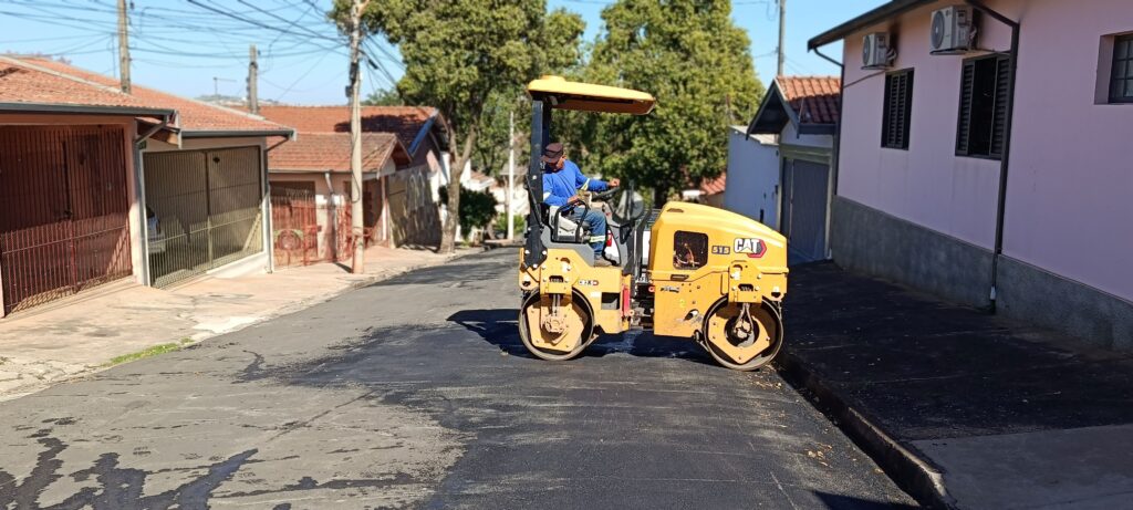 Nesta imagem, vemos um rolo compactador amarelo da marca CAT sendo operado em uma rua residencial que está passando por recapeamento asfáltico. Aqui estão os pontos principais: O Equipamento: É um rolo compactador de solo, modelo CB2.5, que está nivelando e compactando o asfalto novo na via. O Operador: Um homem está conduzindo o maquinário, vestindo uma camisa de manga longa azul e um boné. O Cenário: A cena se passa em uma rua inclinada em um bairro residencial. O asfalto novo, escuro e liso, contrasta com o restante da via. Ao fundo e nas laterais, é possível ver casas típicas, árvores e postes de iluminação, indicando um dia ensolarado.