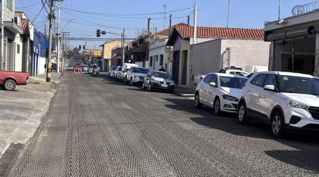Rua em leve aclive vista em perspectiva, com o asfalto recém‑processado (padrão de fresagem) ocupando a maior parte do primeiro plano. Pavimento: camada fresada com textura em malha (sua superfície mostra sulcos e relevo típicos antes da aplicação do novo asfalto). Tráfego e veículos: fila de carros estacionados à direita da via, em sua maioria brancos; ao fundo, alguns veículos em movimento e uma motocicleta se aproximando. Obras/segurança: cones ou sinalização laranja visível ao longe, indicando trabalho na pista. Calçadas e fachadas: prédios baixos comerciais/residenciais alinhados em ambos os lados — fachadas pintadas em tons claros e algumas portas metálicas fechadas; uma caminhonete vermelha estacionada à esquerda. Infraestrutura: vários postes com fiação aérea cruzando a cena; semáforos visíveis ao fundo na interseção. Céu e luz: dia claro, céu azul sem nuvens; sombras suaves indicam iluminação solar direta. Sensação geral: cena urbana cotidiana durante manutenção viária, organizada e em andamento, com predominância de tons neutros e presença clara de atividade de rua.