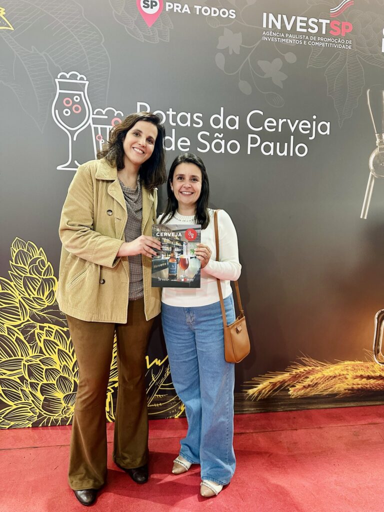 Duas mulheres sorrindo posam lado a lado em frente a um painel promocional. Elas seguram juntas um folheto com imagens de cervejas; o título no fundo diz “Rotas da Cerveja de São Paulo” e há o logo “INVESTSP” no canto superior direito, indicando um estande ou evento relacionado à promoção e turismo cervejeiro. Mulher à esquerda: cabelos castanhos escuros na altura dos ombros, casaco de veludo cotelê bege, blusa estampada por baixo e calça marrom. Usa sapatos pretos. Mulher à direita: cabelos lisos escuros, blusa branca de manga comprida, jeans azul claro e bolsa transversal marrom. Sapatos nude com detalhe claro. Cenário: parede escura com ilustrações relacionadas à cerveja (copos, lúpulo e espigas), chão com carpete vermelho — atmosfera de feira, exposição ou lançamento. Expressão e postura: ambas parecem descontraídas e felizes, posando de forma amigável para a foto.