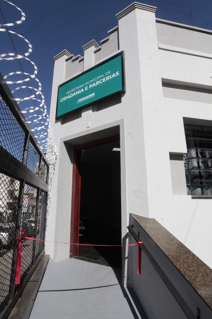 Esta foto mostra a fachada de um prédio público — a Secretaria Municipal de Cidadania e Parcerias — no momento de uma inauguração ou liberação de acesso. Placa verde acima da entrada com o texto “SECRETARIA MUNICIPAL DE CIDADANIA E PARCERIAS” e o logo de Piracicaba. Fachada branca, de linhas retas e acabamento simples; janela com grades à direita. Entrada com porta de madeira aberta, interior escuro visível. Rampa de acesso ao prédio, com corrimão à direita. Fita vermelha esticada bloqueando a passagem na entrada/rampa, indicando cerimônia de corte de fita. Grade metálica à esquerda protegida por concertina (fio farpado em espiral) no topo. Céu azul e sombras fortes, sugerindo luz solar intensa e dia claro.