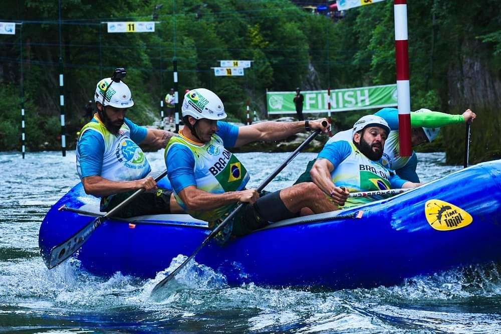 Esta imagem mostra uma equipe brasileira de rafting durante uma competição em um percurso de águas bravas. Aqui estão os detalhes da cena: O bote: Eles estão remando em um bote inflável azul brilhante. Na lateral do bote, há um adesivo amarelo escrito "TIME TRIAL", indicando que é uma prova de contrarrelógio. A equipe: São quatro atletas no bote, todos vestindo uniformes idênticos: camisas brancas com detalhes em azul e verde (cores da bandeira do Brasil), capacetes brancos com o brasão da bandeira brasileira e coletes de proteção também nas cores do Brasil. A ação: Eles estão concentrados e remando com força. O atleta que está à frente, do lado direito, está passando por um poste de sinalização da prova (vermelho e branco), o que sugere que estão navegando um percurso técnico com obstáculos. O ambiente: O cenário é um rio cercado por mata, com marcas de balizamento numeradas penduradas acima da água e uma placa verde ao fundo escrita "FINISH", indicando que estão próximos ao final do percurso. O movimento da água em volta do bote mostra a velocidade e a intensidade da correnteza.