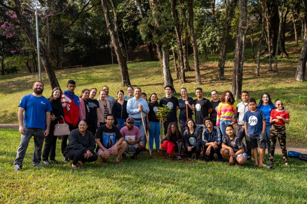 Esta é uma bela foto de grupo ao ar livre, em um ambiente que parece ser um parque ou área verde, celebrando uma ação de plantio. Aqui estão os detalhes: O Grupo: Há um grande número de pessoas, variando entre jovens, adolescentes e adultos, todas reunidas para uma foto coletiva. Todos parecem estar em um momento descontraído e positivo. A Ação: No centro do grupo, é possível ver uma pequena árvore recém-plantada. Um homem, que parece ser um dos organizadores ou um professor, segura uma pá, indicando que eles acabaram de realizar o plantio. O Cenário: O fundo é uma área verde muito bonita, com árvores altas que criam sombras suaves no gramado. É um dia ensolarado e agradável, ideal para atividades ao ar livre. Atmosfera: A cena transmite um forte espírito de comunidade, união e cuidado com o meio ambiente. Muitos estão sorrindo, e o grupo parece bem integrado. É uma imagem que registra um momento muito especial de cidadania e preservação ambiental!