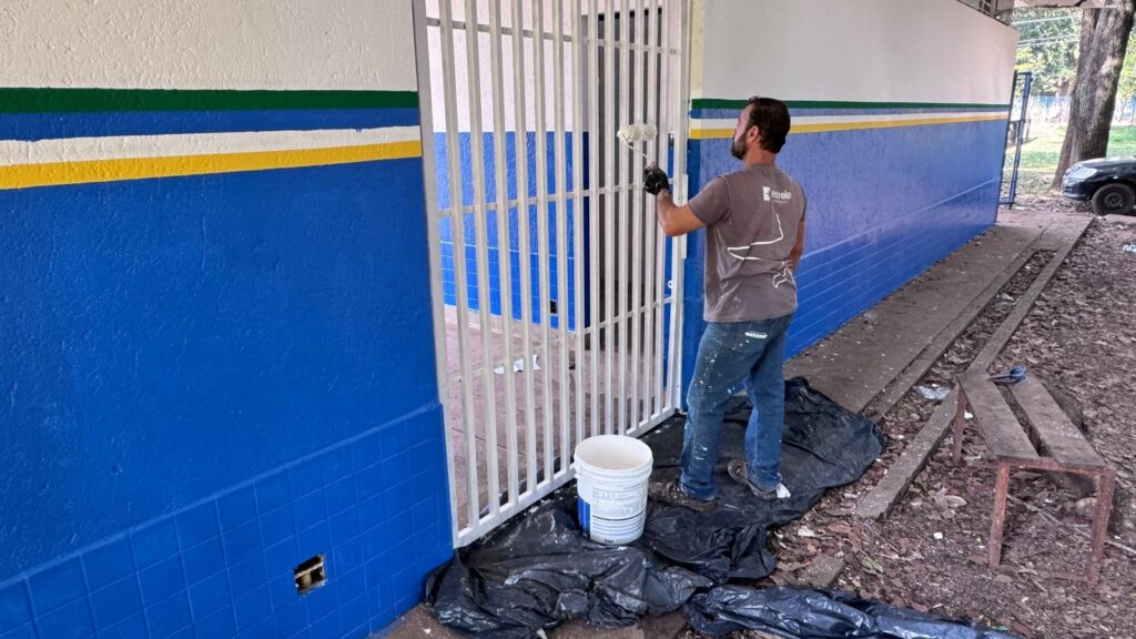 A imagem mostra um homem pintando um portão de grades verticais brancas com um pequeno rolo. Ele usa uma camiseta cinza com a palavra "estrela" e calças jeans manchadas de tinta. O portão está fixado em uma parede pintada de azul, branco, verde e amarelo. Há um balde de tinta branco no chão, sobre uma lona preta que protege a área do solo, onde também se vê um banco de madeira rústico. O ambiente parece ser externo, com árvores e um carro ao fundo à direita.