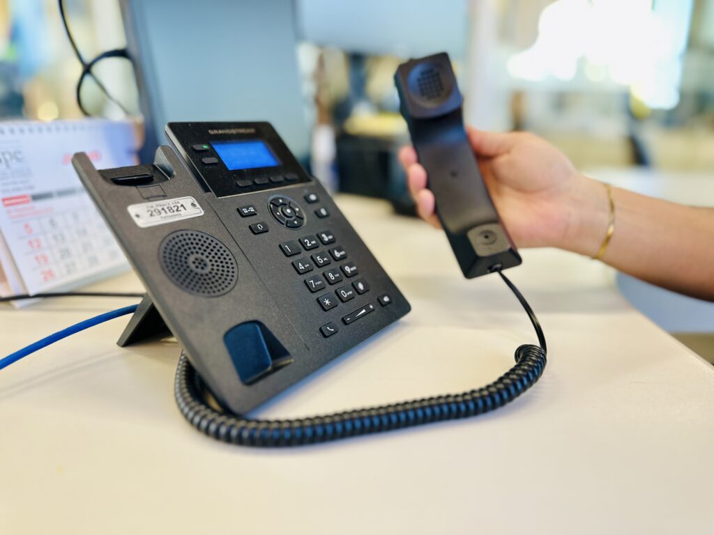 A foto mostra um telefone IP de escritório sobre uma mesa, em um ambiente de trabalho. O Telefone: É um aparelho preto da marca "Grandstream", com teclado numérico padrão, uma tela pequena iluminada em azul e um monofone. Nota-se uma etiqueta de patrimônio colada na lateral do aparelho, indicando "P.M. PIRACICABA 291821". A Ação: Uma mão segura o monofone do telefone, levantando-o da base, como se a pessoa estivesse prestes a atender uma chamada ou discar um número. A pessoa usa uma pulseira dourada fina no pulso. O Ambiente: O cenário é uma mesa de escritório clara. Ao fundo, à esquerda, é possível ver parte de um calendário de mesa. O foco da foto está nítido no telefone, enquanto o fundo apresenta um desfoque suave (efeito bokeh), típico de ambientes de escritório bem iluminados.