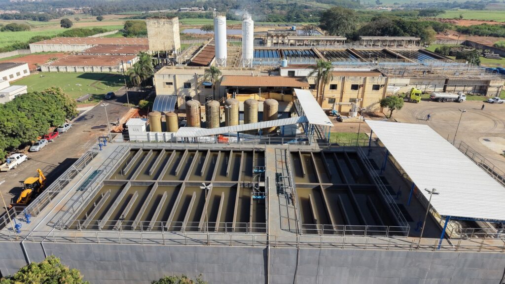 Esta é uma vista aérea ampla de uma Estação de Tratamento de Água (ETA). A estrutura é bastante completa e organizada em diferentes setores de processamento: Tanques de Sedimentação/Floculação: Em primeiro plano, vemos grandes tanques retangulares divididos por várias paredes internas verticais, onde a água passa pelo processo de tratamento. O nível da água é visível e tem um tom acastanhado. Instalações Principais: Atrás dos tanques, há um conjunto de prédios administrativos e operacionais de cor bege, com telhados de telhas cerâmicas e metálicas. Destacam-se dois grandes silos verticais brancos com a marca "SEMAE" estampada, utilizados para armazenamento de produtos químicos. Equipamentos: Entre os prédios e os tanques, há fileiras de tanques cilíndricos maiores (aparentemente filtros ou reatores). Entorno: Ao redor da planta, há uma área pavimentada onde se vê um caminhão amarelo e outros veículos estacionados. O fundo da imagem mostra campos verdes, vegetação e áreas de cultivo, indicando que a estação está situada em uma região mais afastada ou rural. A cena é banhada pela luz do sol, o que sugere um dia claro.