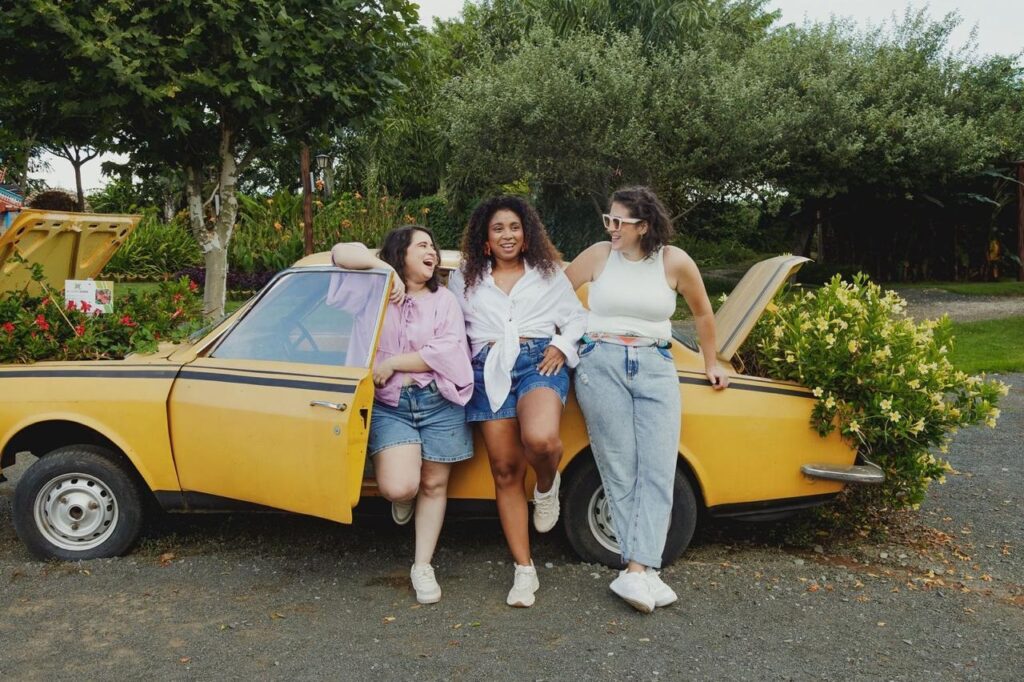 Três mulheres encostadas num carro amarelo antigo em um cenário ao ar livre — descrição detalhada: Composição geral: carro clássico amarelo, com as portas e o porta-malas abertos; as três ocupam a lateral do veículo, alinhadas e próximas, formando um grupo unido e descontraído. As pessoas (sem identificar): À esquerda: mulher com camiseta rosa clara de mangas soltas e shorts jeans curtos; apoia o braço sobre a porta aberta e ri olhando para o centro. Ao centro: mulher de pele mais escura, camisa branca amarrada na cintura e shorts jeans; senta parcialmente na beirada do carro, com expressão sorridente e postura confiante. À direita: mulher usando regata branca e jeans largos de cintura alta; usa óculos de sol claros e apoia a mão no porta-malas levantado, também sorrindo. Roupas e calçados: visual casual e veranil — shorts e jeans, camisetas leves, tênis brancos em todos os três; cores suaves que combinam com a vibração alegre da cena. Carro e detalhes: veículo retro amarelo com faixa preta lateral; porta-malas e capô abertos, e plantas/vegetação saindo pelo porta-malas esquerdo e pela traseira direita, dando toque lúdico e quase surreal (parece uso decorativo). Ambiente: área gramada com árvores e arbustos floridos ao fundo; chão de cascalho à frente, atmosfera de parque ou jardim bem cuidado. Iluminação e clima: luz natural suave — dia claro, clima agradável; cores vibrantes e sombra leve indicam fim de manhã ou tarde. Atmosfera e narrativa: sensação alegre, de amizade e espontaneidade — risos, descontração e cumplicidade; a presença das plantas no carro adiciona um tom criativo e bucólico à cena. Sentimento visual: fotografia calorosa, otimista e estilosa, perfeita para campanha de lifestyle, ensaio informal ou registro de passeio entre amigas.