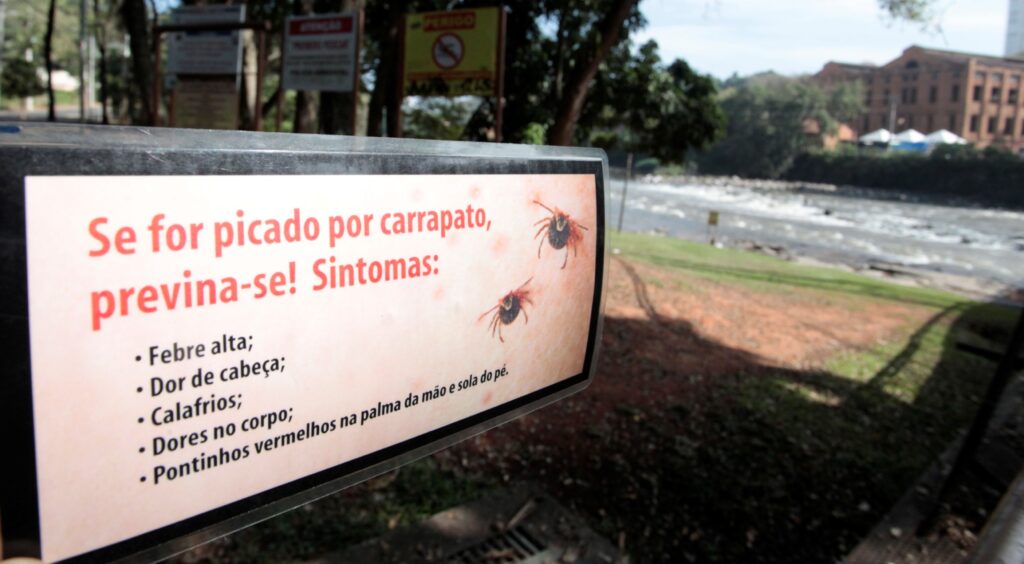 Placa de aviso em primeiro plano indicando prevenção contra picada de carrapato; ao fundo, um parque à beira de um rio. Placa: ocupa a parte esquerda/frontal da imagem, com texto grande em vermelho “Se for picado por carrapato, previna‑se! Sintomas:” e lista em bullets (febre alta, dor de cabeça, calafrios etc.). Há ilustrações de carrapatos ao lado do texto. Ambiente próximo: faixa de grama e pequena área de terra entre a placa e o rio; sombras de árvores sobre o chão. Rio: fluxo visível, com corredeiras suaves e margem de pedras; água clara refletindo o céu. Fundo: árvores e um prédio grande de alvenaria (aparentemente antigo ou industrial) do outro lado do rio; também surgem toldos brancos e mais vegetação. Iluminação/clima: dia claro, luz forte e sombras nítidas — sensação de tarde ensolarada. Atmosfera: área pública ao ar livre, aviso de saúde voltado a quem circula pela vegetação/rio para alertar sobre riscos de carrapatos.