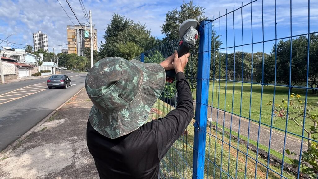 Nesta imagem, vemos uma pessoa de costas trabalhando ao ar livre. Ela está usando um chapéu de estilo "pescador" com estampa camuflada e uma camisa de mangas compridas preta. A pessoa está segurando uma esmerilhadeira (ou serra elétrica portátil) e parece estar cortando ou ajustando o topo de um poste metálico que faz parte de uma cerca de tela azul. O cenário ao redor mostra uma rua pavimentada à esquerda, com um carro passando ao longe e alguns prédios, enquanto à direita há uma área verde, possivelmente um parque ou praça, com gramado e árvores. O céu está parcialmente nublado.