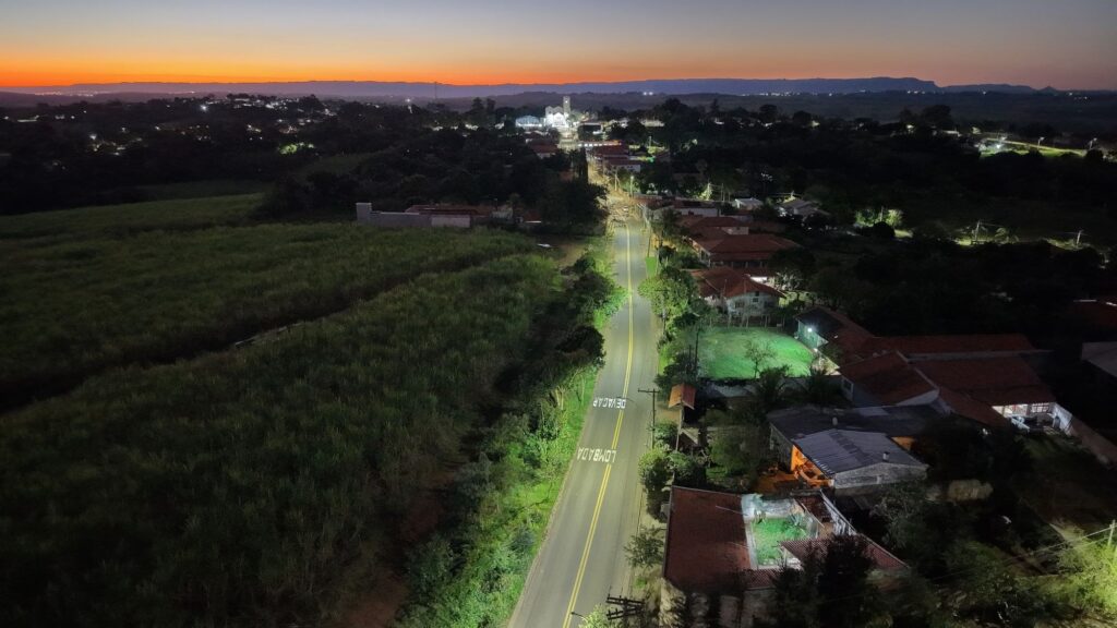Foto aérea panorâmica ao entardecer/crepúsculo, mostrando uma estrada reta que leva a um pequeno povoado. Horizonte com faixa laranja/âmbar acima de morros escurecidos destaca o fim do dia. Estrada e sinalização Via de pista dupla com duas linhas amarelas centrais. Pintura no asfalto com as palavras “DEVAGAR” e “LOMBADA” visíveis em sentido contrário ao ponto de captura. Iluminação pública ao longo da via que cria manchas de luz verdes/amareladas sobre a pista e calçadas. Povoado e construções Casas com telhados de cerâmica avermelhada agrupadas ao lado direito da estrada; algumas residências têm quintal iluminado por holofotes e janelas acesas. Ao fundo, núcleo mais iluminado com construção maior — possivelmente igreja ou prédio público — que funciona como ponto focal luminoso na cena noturna. Pouca movimentação visível; ambiente tranquilo. Vegetação e entorno À esquerda da estrada grande área de vegetação/plantação escura, contrastando com a faixa iluminada da rua. Muitas árvores e trechos de mata entre casas e no horizonte, conferindo ar semi-rural à paisagem. Iluminação, cores e clima Céu em degradê: tons quentes perto do horizonte (laranja) evoluindo para azul escuro no topo; contrasta com as luzes frias e pontuais do vilarejo. Atmosfera calma e serena, sensação de fim de dia com começo da iluminação noturna. Composição e enquadramento Perspectiva aérea/alto — possibilita perceber a relação entre estrada, campo e povoado; linha da estrada guia o olhar do primeiro plano até o ponto luminoso no fundo. Boa profundidade: primeiro plano detalhado (casas e postes), meio com vegetação e estrada, fundo com morros e céu colorido.