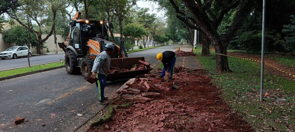 Esta é uma cena de trabalho urbano de manutenção ou obras públicas na calçada. Ação: Dois trabalhadores, equipados com capacetes de segurança e uniformes, estão removendo pedaços de pavimento (parece ser concreto ou pedras antigas) da calçada. Eles estão usando uma retroescavadeira (marca Case 580N) para auxiliar no serviço; a caçamba da máquina está cheia de entulho. O Ambiente: É uma via arborizada e tranquila. Há uma rua pavimentada ao lado, com um carro branco estacionado. O local está sinalizado com fita zebrada (amarela e preta) para isolar a área de trabalho. Detalhes: O solo exposto é de terra vermelha, típica de muitas regiões do Brasil. A iluminação sugere um dia nublado ou final de tarde. Parece ser um serviço de reparo na infraestrutura da calçada, provavelmente realizado por uma empresa de saneamento ou pela prefeitura local.