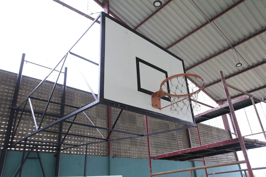 Quadra coberta com foco em uma tabela de basquete vista de baixo e em close médio. Elementos principais: tabela retangular branca com contorno preto e aro laranja com rede de corda parcialmente desgastada. Estrutura: suporte metálico preto que sustenta a tabela, formando um treliçado; andaime/escada metálica vermelha ao lado, provavelmente usado para manutenção. Ambiente: cobertura com telhas metálicas e vigas vermelhas aparentes; parede lateral em blocos de cimento vazados e pintura turquesa na parte inferior. Iluminação: luz natural forte entrando pelas aberturas laterais, criando alto contraste (céu muito claro ao fundo) e sombras suaves dentro da quadra. Estado: equipamentos com sinais de uso (tinta descascada no andaime, rede e aro não novos), mas funcionais; cena transmite um espaço esportivo simples, possivelmente em escola ou ginásio comunitário.