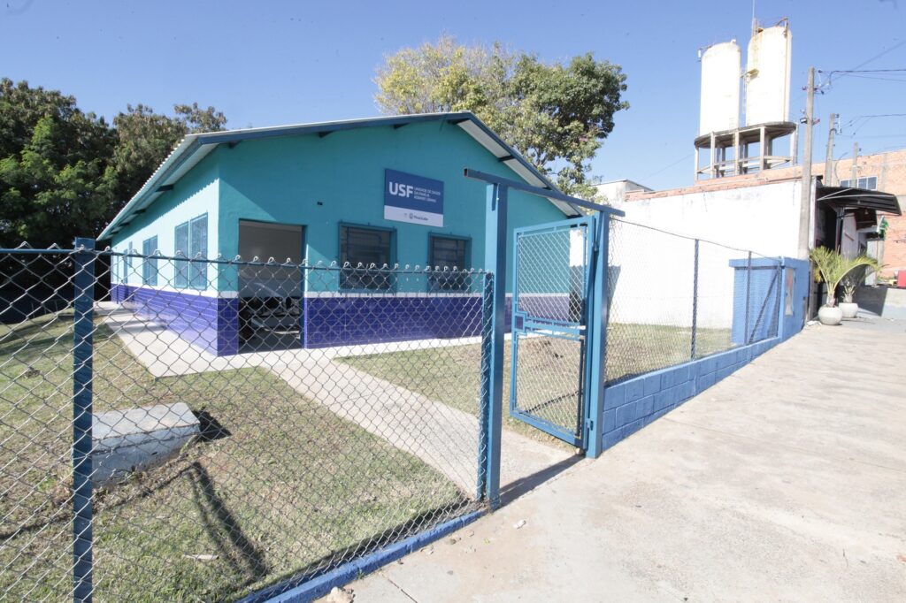 Esta é uma imagem de uma Unidade de Saúde da Família (USF). Edifício: O prédio é de alvenaria, pintado com uma cor turquesa na parte superior e um revestimento de pastilhas azuis escuras na parte inferior das paredes. Identificação: Na fachada, há uma placa branca com o logotipo e a inscrição "USF - Unidade de Saúde da Família Kobayat Libano", além do brasão da prefeitura de Piracicaba. Acesso e Entorno: O prédio é cercado por uma cerca de alambrado com estrutura metálica azul. Há um portão aberto que dá acesso a uma calçada de concreto que leva à entrada principal. Detalhes Adicionais: O dia está ensolarado e com céu azul limpo. Ao fundo, do lado direito, observa-se uma estrutura elevada com dois grandes silos brancos (caixas d'água ou reservatórios). O terreno possui uma área gramada na frente e uma rampa de acesso pavimentada na lateral direita.