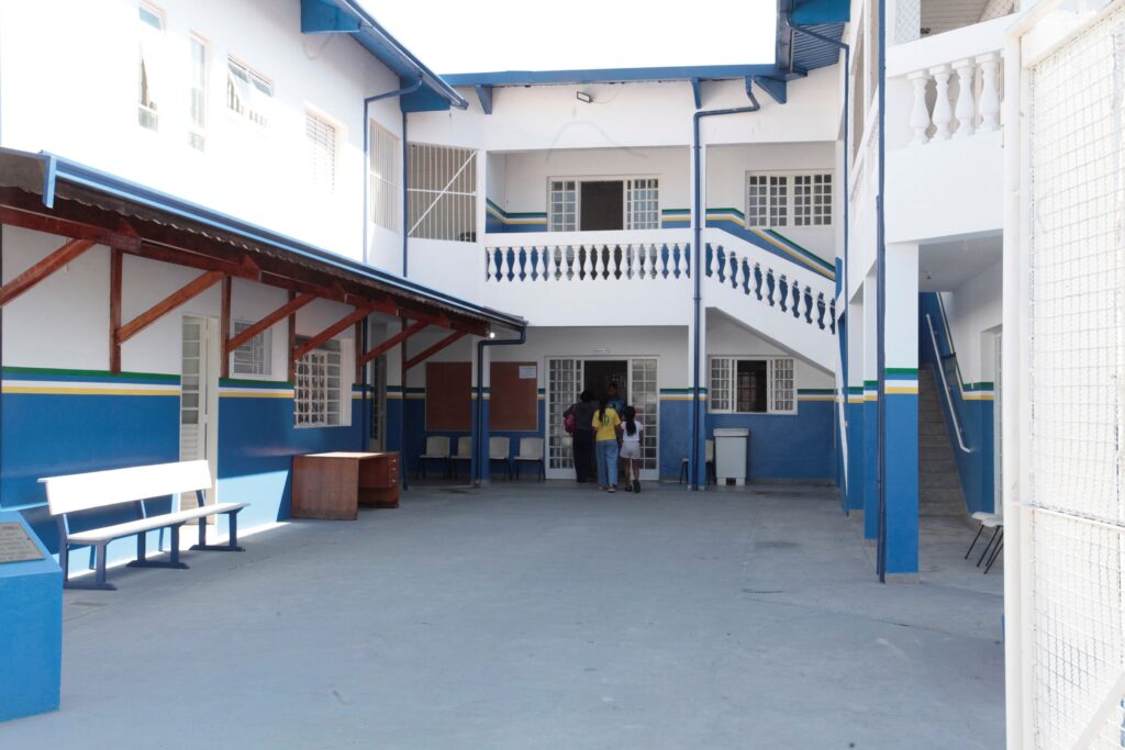Pátio interno de uma escola ou instituição, visto em dia claro. Arquitetura e cores: prédio de dois pavimentos pintado em branco e azul com faixas horizontais verde e amarela; grades nas janelas e guarda-corpo com balaústres brancos na parte superior. Telhado com beirais azuis e estrutura de madeira aparente em um corredor lateral. Área e mobiliário: piso cimentado amplo, bancos e cadeiras ao longo das paredes, um balcão/armário de madeira e uma lixeira branca perto da entrada. Acessos: escada à direita conduz ao segundo andar; passagem central com portas de grade que levam a salas internas. Pessoas: um pequeno grupo (adultos e uma criança) entrando pela porta central, vistos de costas — não é possível identificar quem são. Iluminação e sensação: luz natural forte, sombras suaves; ambiente organizado e tranquilo, típico de escola em horário de atividade.