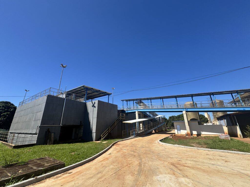 Esta imagem mostra o que parece ser uma estação de tratamento de água ou uma instalação industrial semelhante. Estrutura principal: À esquerda, destaca-se uma grande construção de concreto cinza, com escadas metálicas e cercas de proteção no topo. Passarela: Uma longa passarela elevada, pintada de azul, conecta diferentes partes da planta, permitindo a circulação entre as estruturas. Solo: Um caminho de terra batida (ou concreto com bastante terra acumulada) percorre a área, fazendo uma curva que leva a outras instalações ao fundo. Cenário: O dia está com um céu azul muito claro e sem nuvens, o que dá um aspecto bem aberto e ensolarado ao local. Parece um ambiente de trabalho industrial bem funcional e organizado.