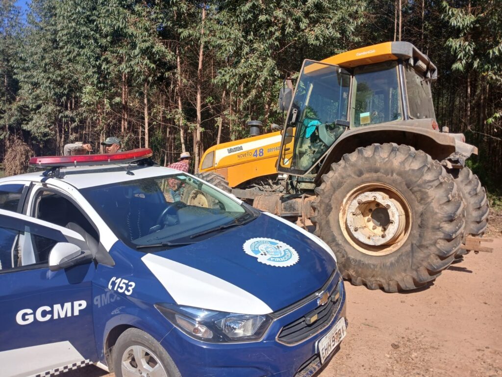 Foto externa em estrada de terra mostrando uma viatura policial municipal à esquerda em primeiro plano e um trator agrícola grande à direita, com área de mata ao fundo. Veículos: na frente, uma viatura azul e branca com a sigla "GCMP" visível na lateral, giroflex vermelho sobre o teto e o brasão da prefeitura/guarda no capô; à direita, um trator amarelo-alaranjado de grande porte (cabine fechada, pneus traseiros muito altos e largos) com marcações e números na carroceria. Pessoas: pelo menos duas pessoas ao fundo, entre a viatura e o trator, em pé junto ao veículo agrícola — não faço identificação nem suposições sobre quem sejam. Ambiente: estrada de terra batida, solo seco com marcas de pneus; faixa de vegetação densa atrás, provavelmente eucaliptos ou árvores altas alinhadas formando um trecho de mata. Condição e detalhes: o trator está visualmente em uso/operacional (sujeira nas rodas e carroceria); a porta da cabine do trator parece aberta; a viatura tem a porta dianteira também aberta, mostrando parte do interior. Iluminação e clima: dia claro com sol forte — sombras definidas e cores saturadas indicando boa luminosidade. Atmosfera: cena de operação ou fiscalização em área rural, com interação entre veículo oficial e máquina agrícola em via não pavimentada.