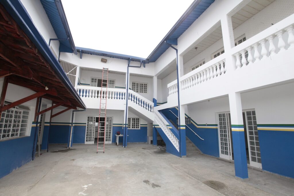 Esta é uma foto do pátio interno de um prédio que parece ser uma escola ou um centro de atendimento público. Aqui estão os detalhes: Arquitetura: O edifício tem dois andares, com paredes brancas e detalhes pintados de azul na base, formando uma faixa decorativa com listras finas em verde e amarelo. Estrutura: Há uma escada externa de concreto com balaustradas brancas que leva ao segundo andar, onde se vê uma varanda aberta com grades de proteção. Cobertura: Uma parte da lateral esquerda possui um telhado com estrutura de madeira aparente. Elementos: Há uma escada de metal longa encostada na parede central, sugerindo algum tipo de manutenção ou trabalho em andamento. Ambiente: O pátio é pavimentado com cimento liso e o local parece ser bem arejado e iluminado pela luz do dia. O lugar tem um aspecto bastante organizado e funcional!