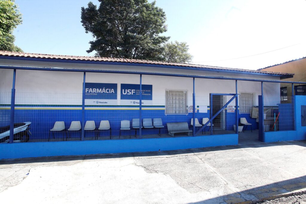 Fachada de uma unidade de saúde pública (farmácia/USF) vista de frente, com grade baixa na parte externa e fileira de cadeiras para espera. Predomínio das cores branco e azul na pintura; faixa decorativa com listras nas cores verde, amarela e azul logo acima do revestimento azulado em meia-altura. Dia claro e céu azul sem nuvens visível acima do telhado. Detalhes arquitetônicos e ambiente Telhado de telhas cerâmicas em tom avermelhado, beiral com estrutura simples. Parede branca lisa na parte superior e azulejos/azulejamento azul na parte inferior. Estrutura metálica pintada de azul sustentando a cobertura do alpendre frontal. Grade metálica horizontal na frente que separa a calçada da área de espera; porta gradeada à direita que parece ser a entrada principal. Janelas com grades brancas e venezianas/grade interna. Sinalização e texto Placas azuis com letras brancas: “FARMÁCIA” à esquerda e “USF” (Unidade de Saúde da Família) à direita; abaixo delas menção a “Vila Fátima / Jardim Primavera” e selo/assinatura da prefeitura de Piracicaba. Pequenos cartazes/branc os afixados próximos à porta (detalhes dos textos menores não legíveis à distância da foto). Mobiliário e objetos Fileira de cadeiras plásticas acopladas e individuais em tons claros (branco, bege) para pacientes na área externa. Um banco estofado de tom cinza ao lado das cadeiras. À direita, dentro da área, uma cadeira e alguns objetos de limpeza encostados (vassouras/rodo). À esquerda, parcialmente visível, a traseira de uma motocicleta estacionada junto à grade. Vegetação e entorno Árvores altas ao fundo, por trás do prédio, com copa densa. Lado direito mostra parte de outro imóvel amarelo ao fundo. Impressão geral / tom da cena Local transmite funcionalidade e organização típica de uma pequena unidade de saúde municipal: cuidado básico, área de espera externa simples e sinalização clara. Atmosfera tranquila, sem pessoas no momento da foto.