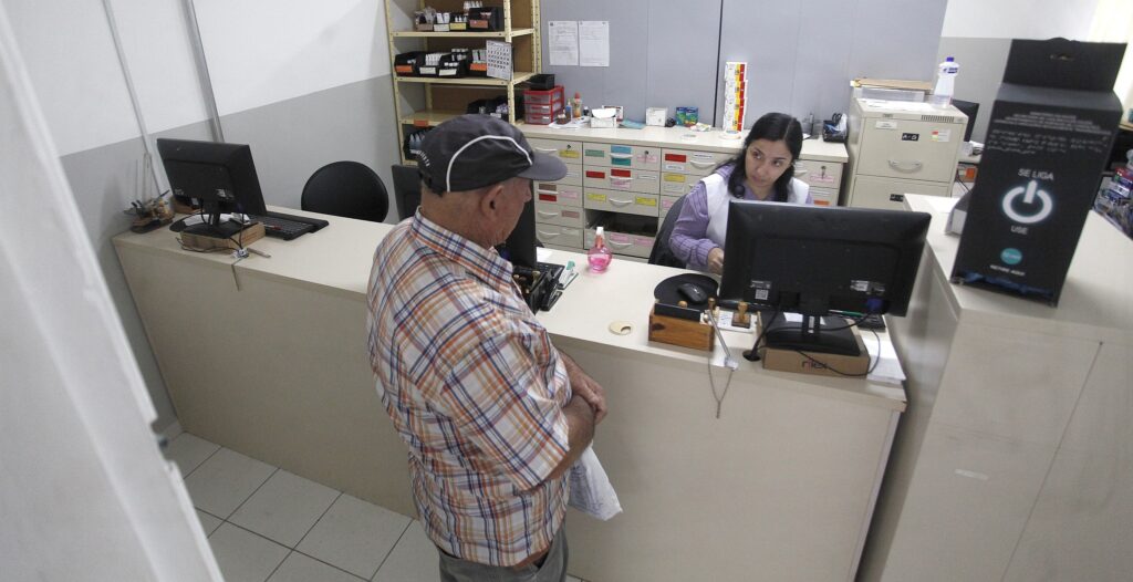 Área de atendimento/recepção de uma unidade (provavelmente de saúde ou farmácia), vista em ângulo levemente superior. Balcão claro separando o atendente do público; ambiente limpo e funcional. Pessoas e ação Homem idoso em primeiro plano, de costas/parcialmente de perfil, usando camisa xadrez em tons de bege/laranja e boné escuro; segura um saco plástico branco com a mão direita apoiada no balcão. Atendente (mulher) sentada atrás do balcão, usando jaleco branco sobre blusa lilás, olhando para o homem e com uma das mãos próxima ao teclado ou ao documento — parece registrar ou conferir algo no computador. Mobiliário e objetos Dois monitores de computador sobre o balcão (um voltado para o atendente, outro mais à esquerda), teclado e mouse. Carimbo(s) e estojo de carimbos em madeira sobre o balcão, fita adesiva, um frasco pequeno rosa (talvez borrifador) e alguns papéis. Ao fundo, armário com muitos gaveteiros identificados por etiquetas coloridas; prateleiras com caixas e material de escritório/medicamentos. Arquivo/arquivo metálico e outros pequenos itens organizacionais ao fundo à direita. Elementos do ambiente Paredes claras com faixa cinza na metade inferior, piso cerâmico claro. Iluminação neutra e ambiente sem outras pessoas visíveis. Tom e impressão Cena cotidiana e rotineira de atendimento: ambiente organizado, tranquilo e funcional, com interação profissional entre usuário e atendente.