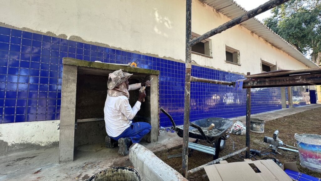 Foto de um canteiro de obra mostrando um trabalhador aplicando acabamento em uma estrutura de concreto à frente de uma parede revestida de azulejos azuis. Composição: vista horizontal ampla, foco na parte central-esquerda onde o operário está agachado dentro de um vão de alvenaria. Trabalhador: homem com camisa branca e calça azul, usando chapéu de abas e proteção no pescoço; ele segura uma talhadeira e martelo, trabalhando o acabamento do vão. Parede: revestimento quase completo com pequenos azulejos quadrados de tom azul vivo cobrindo a metade inferior da fachada; a metade superior está rebocada em massa clara ainda sem pintura. Estrutura/obras: vigas de concreto formando vãos de janelas/portas; andaimes metálicos e pranchas à frente da parede, indicando trabalho em altura e continuidade do revestimento. Ferramentas e materiais: betoneira/rodinha com argamassa, baldes, saco de cimento, peças de madeira e algumas ferramentas espalhadas pelo chão. Ambiente: área externa com chão de terra/grama junto ao prédio; árvores ao fundo à direita, e claridade indicando dia ensolarado com sombras suaves. Estado da obra: acabamento em andamento — azulejos já assentados na parte inferior, resta retoques nos vãos e aplicação final no reboco superior.