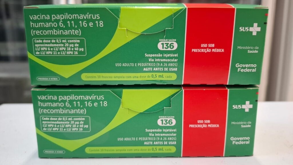 Foto de duas caixas empilhadas de vacina contra o papilomavírus humano (HPV). Aparência geral: duas caixas verdes com detalhes em vermelho e amarelo, alinhadas uma sobre a outra sobre uma superfície branca. Texto visível: “vacina papilomavírus humano 6, 11, 16 e 18 (recombinante)”; informação sobre dose de 0,5 mL; indicação de uso intramuscular; “Uso sob prescrição médica”; selo/SIGLA “SUS”, “Ministério da Saúde” e “Governo Federal”. Informação adicional legível: indicação etária (uso adulto e pediátrico, 9 a 26 anos), instrução “agite antes de usar” e telefone de saúde “Disque Saúde 136”. Condição e contexto: embalagens parecem novas e lacradas; foto tirada em ambiente bem iluminado, com foco nas laterais das caixas.