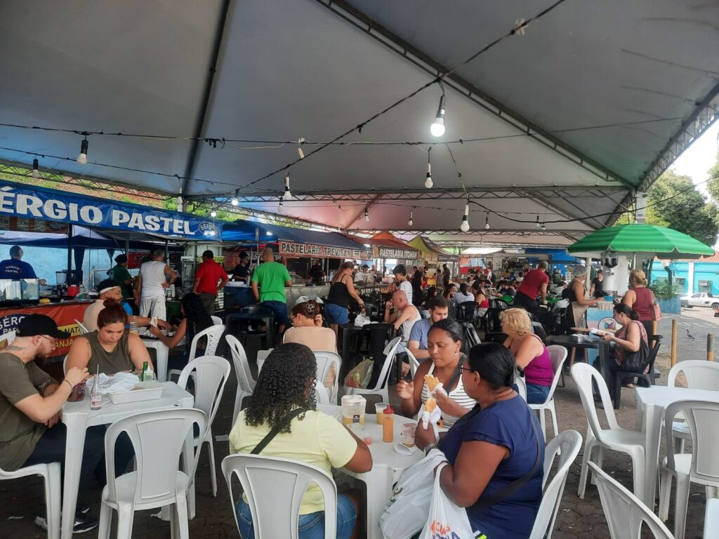 A imagem mostra uma praça de alimentação ao ar livre, instalada sob grandes tendas brancas. O local está movimentado, com várias pessoas sentadas em mesas e cadeiras de plástico branco, comendo e conversando. Em primeiro plano, destacam-se alguns clientes, como duas mulheres à direita que estão comendo e um homem de boné à esquerda. Ao fundo, vê-se uma fileira de barracas de comida, com letreiros como "Sérgio Pastel" e "Pastelaria Trevisan", onde funcionários trabalham no preparo dos alimentos. O ambiente é iluminado por lâmpadas penduradas na estrutura da tenda, e à direita há um guarda-sol verde e branco listrado.