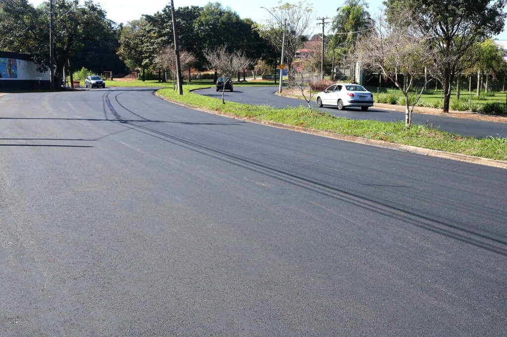 Nesta foto, vê-se uma rua que acabou de passar por um serviço de pavimentação, com asfalto novo, escuro e bem liso. Aqui estão os detalhes: A Rua: A via apresenta uma curva suave e é ladeada por canteiros centrais com grama verde. O asfalto está impecável, sem remendos, cobrindo toda a extensão visível da rua. Trânsito: Alguns carros aparecem circulando pela via, indicando que o tráfego já foi liberado ou está fluindo normalmente. Um carro de cor clara está à direita, e outro veículo (mais escuro) segue ao longe. Ambiente: O local é arborizado, com árvores de porte médio em ambos os lados e nos canteiros, criando um visual bem cuidado. O dia parece estar ensolarado, com sombras marcadas no chão, sugerindo um clima agradável. Ao fundo, percebe-se que é uma área residencial ou de condomínio, com cercas e sinalização de trânsito.