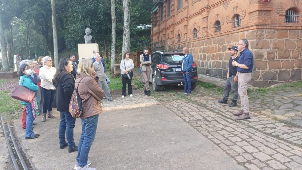 Foto de um pequeno grupo participando de uma reunião ou visita guiada ao ar livre, junto a um edifício histórico de alvenaria. Composição: cerca de 15–20 pessoas dispostas em meia-lua à esquerda da cena, olhando para três pessoas à direita que parecem conduzir a atividade (um fala enquanto os outros escutam). Ambiente e local: prédio de tijolo à vista com base em pedras, janelas gradeadas e escadaria/grade metálica ao fundo; calçamento de paralelepípedo junto a trecho de piso de cimento; vegetação e árvores altas atrás de um busto sobre pedestal. Pessoas: grupo misto de adultos e idosos, maioria vestindo roupas casuais de frio (jaquetas, casacos, botas); uma pessoa usa máscara; bolsas e mochilas visíveis em algumas participantes. Objetos e veículos: um automóvel preto estacionado ao lado do prédio, encostado na área gramada; pequeno cartaz no vidro traseiro do carro (não legível à distância). Ação e atmosfera: postura atenta e tranquila — parece uma palestra, tour histórico, reunião comunitária ou atividade educativa; iluminação suave de final de tarde. Detalhes menores: folhas e flores caídas no chão à esquerda, um busto em pedestal perto das árvores que indica importância histórica do local, e rampa/caldeirão metálico ao fundo.