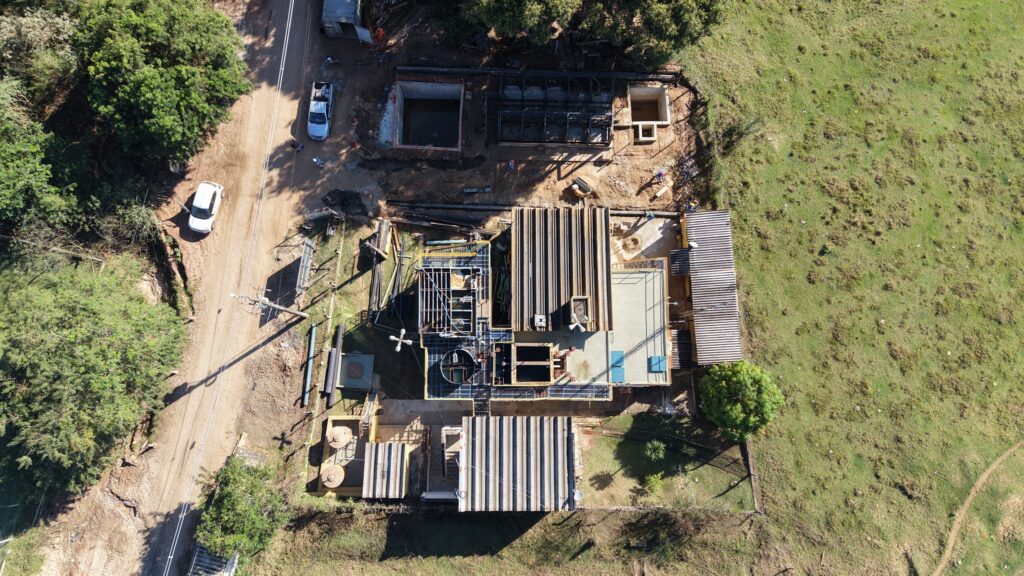 A imagem mostra uma vista aérea de um canteiro de obras em um ambiente rural. Aqui estão os detalhes: Estruturas em Construção: Várias construções em diferentes estágios, com estruturas metálicas e coberturas de telhado visíveis. Algumas áreas têm lajes de concreto já colocadas. Equipamentos e Materiais: Há tubos e vigas de metal espalhados pelo chão, indicando atividade intensa de construção. Tanques e caixas de armazenamento podem ser vistos, possivelmente relacionados ao abastecimento de água ou outros materiais. Veículos: Dois carros estão estacionados em uma estrada de terra ao lado do canteiro, sugerindo acesso fácil ao local. Vegetação: Ao redor, há árvores e áreas gramadas, mostrando um ambiente natural, com o terreno ainda não completamente modificado. Pessoas Trabalhando: Algumas pessoas podem ser vistas em diferentes partes do local, realizando tarefas de construção. Caminho de Acesso: Uma estrada de terra leva ao canteiro, com postes de eletricidade ao longo do caminho. Esse cenário reflete uma fase ativa de construção, com muitos elementos que indicam progresso e desenvolvimento.