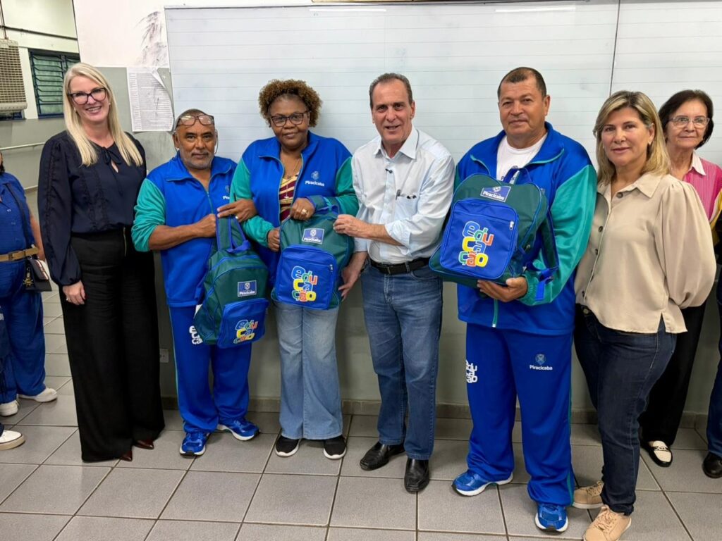 Um grupo de oito pessoas posando em fila dentro de uma sala (provavelmente de escola ou administração), diante de um quadro branco. Todos olham para a câmera e sorriem levemente. Pessoas: sete visíveis frontalmente no centro e à direita, mais uma parcialmente visível à esquerda. Mistura de homens e mulheres adultos; aparentam idades de meia-idade a mais velhos. Roupa e uniformes: três pessoas (dois homens e uma mulher, no centro-esquerda e centro-direita) vestem agasalhos azuis e verdes com a marca “Piracicaba” — parecendo uniforme institucional. Outras usam roupas casuais/formais: camisa social branca (homem no centro), blusa preta longa (mulher à esquerda) e blusa bege (mulher à direita). Objetos principais: três pessoas seguram mochilas azul-esverdeadas com o logotipo colorido “Educação” e um selo com o nome “Piracicaba” — parecem ser kits ou material escolar distribuídos. Ambiente: piso de cerâmica cinza, quadro branco ao fundo e parte de um ar-condicionado/ventilação à esquerda; iluminação interna típica de sala institucional. Composição e impressão: foto formal, possivelmente registrando uma entrega oficial ou evento de distribuição de materiais ligados à educação; clima cordial e institucional.