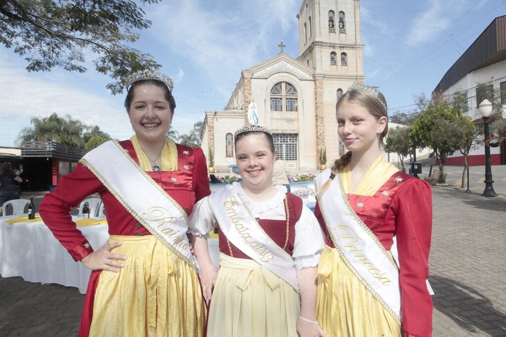 Foto de três jovens em traje cerimonial posando em frente a uma igreja numa praça ensolarada. Observações: Três garotas lado a lado, sorrindo e olhando para a câmera; todas usam tiaras brilhantes. Trajes coordenados: vestidos/saias amarelas com corpetes vermelhos; mangas longas vermelhas e detalhes bordados. Faixas brancas com escrita dourada sobre o peito — parecem títulos festivos (por exemplo: “Rainha”, “Embaixatriz”/“Princesa”), em estilo de festa ou cortejo tradicional. A jovem do centro usa uma faixa e um vestido com babados no colo; parece ser a mais baixa do grupo. Ao fundo há uma igreja com torre e estátua na fachada, árvores, postes e mesas com toalhas e garrafas, sugerindo evento público ou festa local. Iluminação diurna, céu claro com poucas nuvens; sombras suaves no chão indicam sol forte. Atmosfera: comemorativa e formal, possivelmente festival, coroação ou desfile comunitário.
