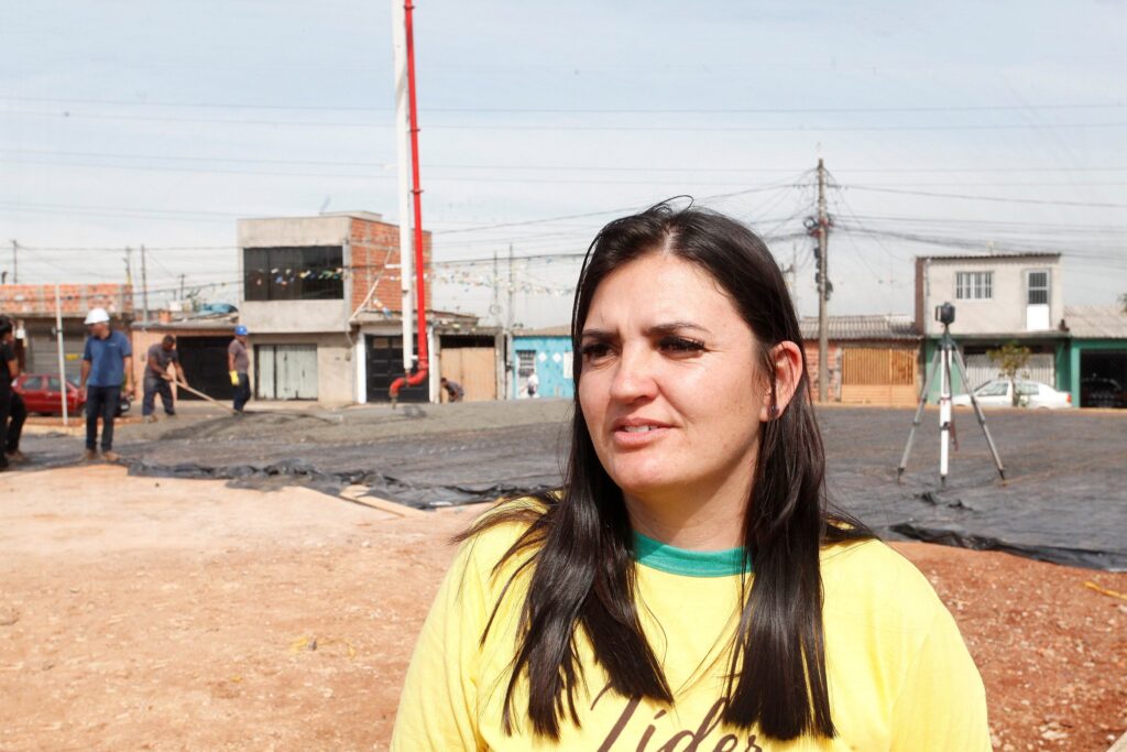 Uma mulher em primeiro plano à direita da foto, vista da altura do peito para cima. Detalhes principais: Aparência: cabelos longos e escuros soltos, pele clara, expressão neutra; usa uma camiseta amarela com gola verde e parte de uma palavra (“Líder”) visível no peito. Enquadramento: ela ocupa a metade direita da imagem, ligeiramente fora do centro; o foco está maior nela, o fundo está em profundidade de campo média. Cena de fundo: área de obra/rua em pavimentação. Ao fundo, trabalhadores usando capacetes e luvas mexem em uma superfície coberta por lona preta e malha de vergalhões — aparenta-se tratar de concretagem ou preparação para concretagem. Equipamentos e elementos: tubo/braço vermelho de bomba de concreto ao centro-esquerda, um tripé com equipamento topográfico à direita ao fundo, caminhões e alguns materiais de construção espalhados. Ambiente urbano: casas simples e prédios baixos ao fundo, fiação aérea visível, céu claro e luz do dia forte, gerando sombras suaves. Atmosfera: sensação de trabalho em andamento e vistoria/fiscalização; a presença da mulher em primeiro plano sugere observação ou entrevista, sem possibilidade de identificação pessoal.
