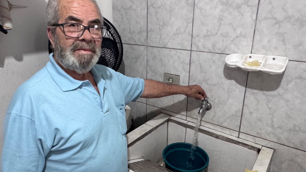 Um homem idoso de óculos azuis e camisa polo está girando a torneira de uma pia, enquanto uma água cai em um balde verde. O homem, que parece ter cerca de 70 anos, tem cabelos grisalhos curtos e uma barba grisalha rala. Ele usa óculos de aro preto e uma camisa polo azul clara de manga curta. Suas mãos estão enrugadas e ele está gentilmente girando a torneira com a mão direita, que está em foco na cena. Seu olhar está direcionado para a câmera, com uma expressão neutra, mas talvez um leve sorriso nos lábios, ou uma expressão de quem está sendo fotografado enquanto realiza uma tarefa. atrás dele, há um ventilador preto, de cujo contorno se vê a grade em espiral. A cena se passa em um ambiente interno, possivelmente um banheiro ou área de serviço, como sugerido pelas paredes revestidas de azulejos brancos com veios cinza e uma pia de concreto com bordas desgastadas. A pia está cheia de água, e um balde verde escuro está posicionado abaixo da torneira para coletar a água que está correndo. Há uma saboneteira branca fixada na parede acima da pia, com o que parecem ser resíduos de sabão dentro dela. Uma tomada elétrica com duas entradas redondas e um pino de aterramento está visível na parede, entre a torneira e o ventilador. A iluminação é geral e uniforme, sem sombras fortes, o que sugere uma luz artificial ou um dia nublado. As cores predominantes são o azul claro da camisa do homem, o branco e cinza dos azulejos e da saboneteira, e o verde escuro do balde. A atmosfera da imagem é cotidiana e simples, capturando um momento comum de uma tarefa doméstica. Não há uma sensação de urgência ou drama; em vez disso, há uma calma e uma normalidade. O humor é pacífico e talvez um pouco melancólico, devido à idade do homem e à simplicidade do cenário.