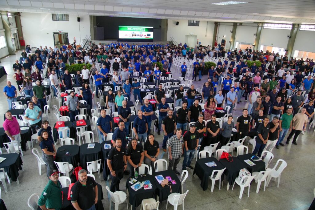 Uma vasta multidão de colaboradores está reunida em um amplo centro de convenções, posando para uma fotografia oficial em um ambiente corporativo organizado, capturando um momento de unidade e confraternização profissional. A composição da imagem é estruturada em uma perspectiva de ângulo elevado que cobre todo o salão, revelando uma organização simétrica de mesas redondas com toalhas pretas e cadeiras brancas. As pessoas estão distribuídas por todo o espaço, desde o primeiro plano até o fundo, onde o palco com uma tela grande serve como ponto focal central. A disposição segue um padrão de preenchimento de espaço, com os participantes ocupando tanto os assentos quanto os corredores, criando uma sensação de grande escala e participação em um evento corporativo de porte significativo. Os sujeitos são um grande grupo diversificado de homens e mulheres de várias idades, apresentando uma ampla gama de etnias e estilos. No primeiro plano, indivíduos estão posicionados de forma mais próxima à câmera, vestindo trajes casuais e uniformes de trabalho, incluindo camisas polo, camisetas e camisas sociais, muitas delas com logotipos de empresas. Suas expressões são variadas, com muitos sorrindo para a câmera ou mantendo posturas atentas, refletindo um comportamento coletivo de colaboração. A vestimenta é predominantemente composta por tons de azul, preto e cores sóbrias, condizente com um ambiente de trabalho industrial ou corporativo. A imagem é uma fotografia digital de alta resolução, tecnicamente executada com uma lente grande angular para abarcar todo o auditório sem distorção excessiva. A nitidez é mantida em profundidade, permitindo que os detalhes dos rostos no primeiro plano e os contornos das pessoas ao fundo sejam discerníveis. O estilo é documental e informativo, focado em registrar a magnitude de um encontro corporativo. A execução destaca o contraste entre as superfícies das mesas escuras e a claridade das cadeiras, contribuindo para a legibilidade visual da cena, mantendo um equilíbrio de tons neutros em todo o quadro. O cenário é um salão industrial amplo com piso de concreto polido e um teto alto com estruturas metálicas visíveis. A iluminação é difusa e uniforme, proveniente de grandes luminárias de teto, que iluminam todo o espaço de forma equilibrada, eliminando sombras fortes e criando uma atmosfera profissional e brilhante. O ambiente é complementado por uma tela no fundo que projeta logotipos corporativos, reforçando o caráter institucional da reunião. A luz natural entra suavemente pelas janelas laterais, contribuindo para a clareza geral da cena e conferindo um tom de transparência e seriedade ao evento, enquanto a organização espacial sugere um ambiente de trabalho bem planejado.