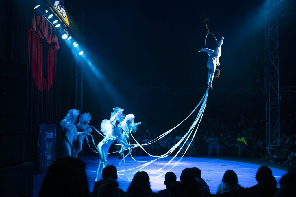 A imagem mostra uma apresentação circense em um palco iluminado por luzes azuis. No alto, uma artista realiza uma acrobacia aérea suspensa em um aro, enquanto longas fitas azuis descem até o chão. Abaixo, outros artistas usam figurinos brilhantes e volumosos, acompanhando a apresentação com as fitas. O público observa sentado ao redor do palco, criando uma atmosfera teatral e encantadora. Ao lado esquerdo, há cortinas vermelhas e estruturas de iluminação.
