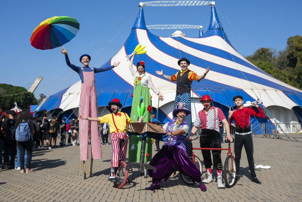 A imagem mostra um grupo animado de artistas de circo em frente a uma grande tenda azul e branca. Eles estão vestidos com roupas coloridas e alegres — alguns em pernas de pau, outros com chapéus e maquiagem de palhaço. Há também um monociclo e um pequeno triciclo, além de objetos de malabarismo e um guarda-chuva multicolorido. O cenário transmite energia, diversão e o espírito vibrante típico de um espetáculo circense.